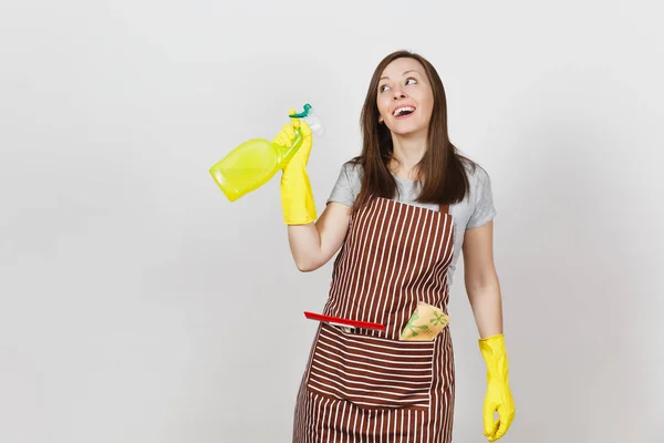 Jovem dona de casa sorridente em luvas amarelas, avental listrado, pano de limpeza, rodo no bolso isolado no fundo branco. Mulher empregada segurando frasco de spray com líquido mais limpo. Espaço de cópia da garrafa . — Fotografia de Stock