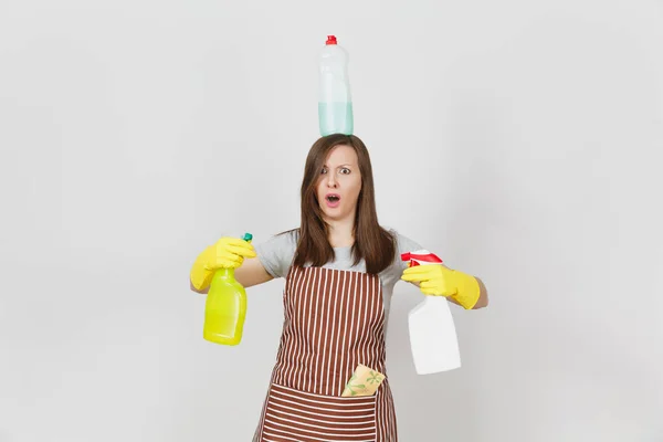 Fun housewife in yellow gloves, striped apron, cleaning rag in pocket on white background. Woman holding in hands, on head bottles with cleaner liquid for washing dishes. Copy space for advertisement. — Stock Photo, Image