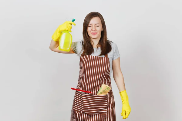 Joven ama de casa divertida en guantes amarillos, delantal rayado, trapo de limpieza, escobilla en bolsillo aislado sobre fondo blanco. La ama de llaves mujer dispara desde la botella de aerosol con líquido más limpio. Espacio de copia de botella . —  Fotos de Stock