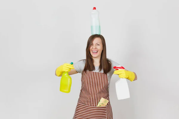 Fun housewife in yellow gloves, striped apron, cleaning rag in pocket on white background. Woman holding in hands, on head bottles with cleaner liquid for washing dishes. Copy space for advertisement. — Stock Photo, Image