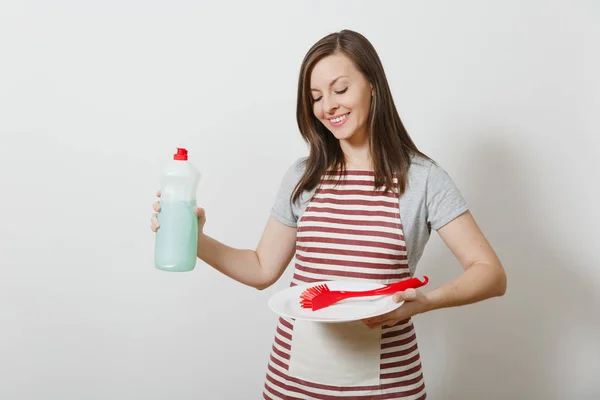 家庭主妇在白色背景条纹围裙隔离。女管家拿着瓶子清洁的液体, 红刷为洗盘子, 白色空心圆盘子。广告复制空间瓶. — 图库照片