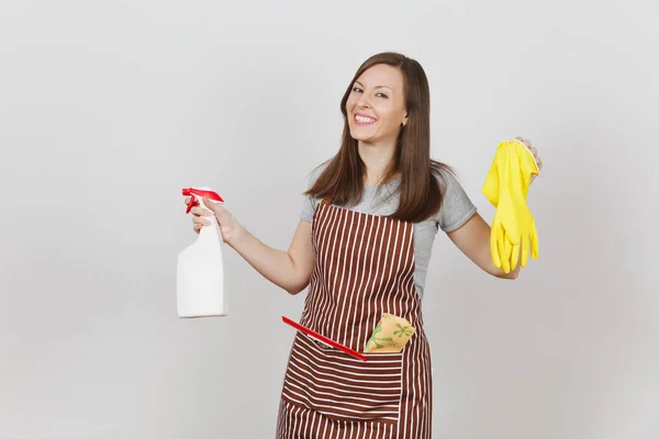 年轻微笑的家庭主妇在条纹围裙与清洁抹布刮在白色背景的口袋隔离。女管家手持喷雾瓶, 用干净的液体黄色手套。瓶拷贝空间. — 图库照片
