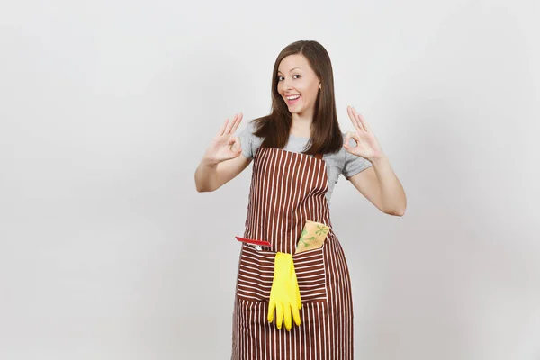 Jeune femme au foyer souriante en tablier rayé avec chiffon nettoyant, gants jaune raclette dans une poche isolée sur fond blanc. Jolie femme de ménage montrant OK geste regardant caméra. Pour la publicité — Photo