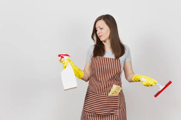 Young sad upset bewildered housewife in yellow gloves striped apron cleaning rag squeegee in pocket isolated on white background. Woman spread hands, hold spray bottle with cleaner liquid. Copy space. — Stock Photo, Image