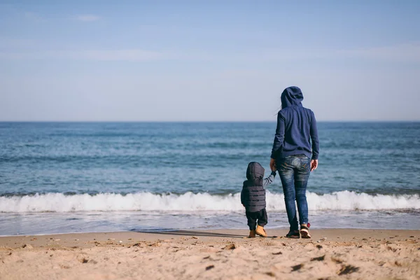 Mujer joven de pie en el fondo de la playa de arena de mar cogido de la mano niño lindo bebé, mirando en el horizonte. Madre, hijito. Día de la familia 15 de mayo, el amor, los padres, los niños concepto. Vista trasera . —  Fotos de Stock