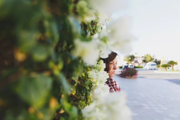 Портрет молодой привлекательной красивой брюнетки, стоящей летом теплый солнечный день в зеленых кустах с белыми цветами перед каменным каменным забором. Чувствительность. Вид сбоку . — стоковое фото