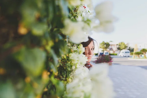 Retrato de joven atractiva hermosa chica morena de pie en verano cálido día soleado en arbustos verdes con flores blancas en frente de valla de roca de piedra. Sensualidad. Vista lateral . — Foto de Stock