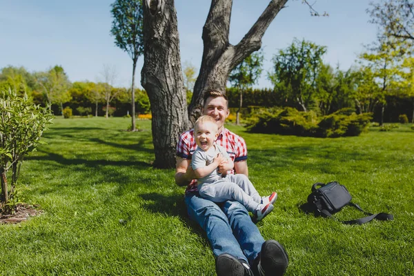 Joyful man walk on green park, rest, have fun, play, high toss up little cute child baby boy sit on grass at tree. Father throw up little kid son. Family day 15 of may, love parents, children concept. — Stock Photo, Image
