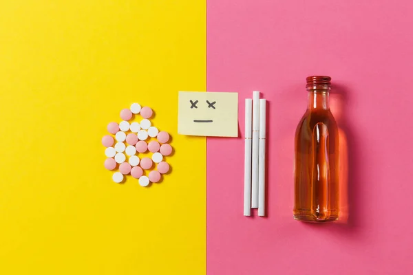 Medicamentos tabletas coloridas píldoras dispuestas abstracto, botella de alcohol, cigarrillos sobre fondo de color rosa amarillo. Papel pegatina cara sonrisa triste. Tratamiento, elección de estilo de vida saludable. Copiar espacio . — Foto de Stock