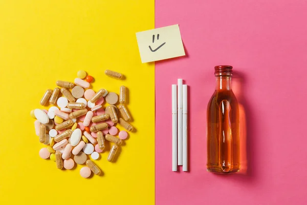 Medicinering färgglada tabletter piller ordnade abstrakt, flaska alkohol, cigaretter på gul rosa ros färgbakgrund. Papper klistermärke leende leende ansikte. Behandling, val hälsosam livsstil. Kopiera utrymme. — Stockfoto