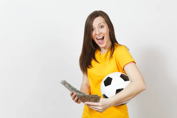 European young fun woman, football fan or player in yellow uniform holding bunch of money banknotes, soccer ball isolated on white background. Sport, play football game, excitement lifestyle concept.