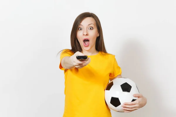 Beautiful European young cheerful woman, football fan or player in yellow uniform hold TV remote, soccer ball isolated on white background. Sport, play football, health, healthy lifestyle concept.