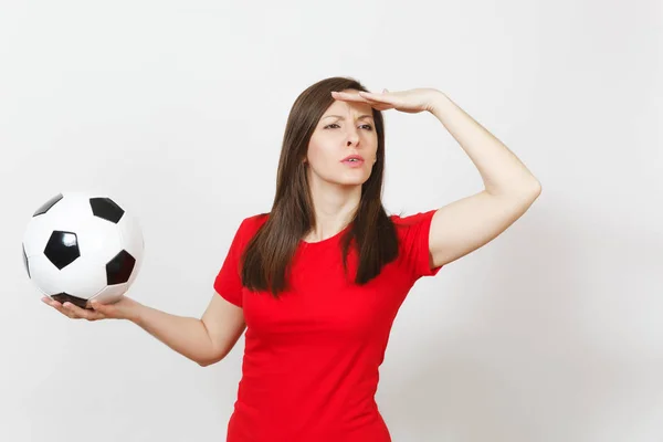 Attractive European young woman, football fan or player in red uniform holding soccer ball, looking into distance isolated on white background. Sport, play football, cheer, healthy lifestyle concept.
