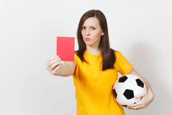 European serious severe young woman, football referee or player in yellow uniform showing red card, holding soccer ball isolated on white background. Sport, play football, healthy lifestyle concept.