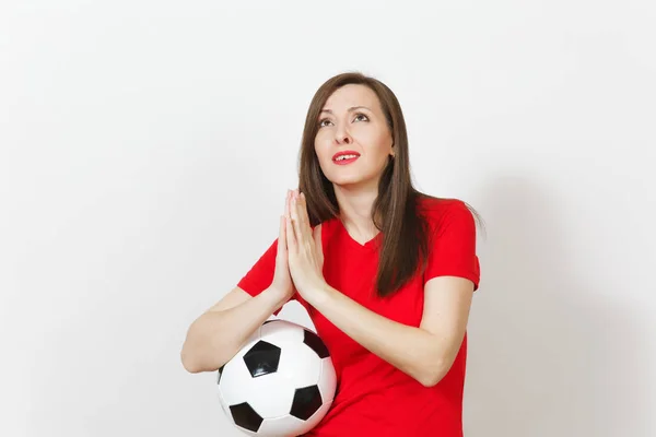 European young woman football fan or player in red uniform folded hands in prayer, hold soccer ball isolated on white background. Sport play football lifestyle concept. Wait special moment. Make wish.