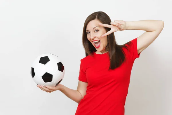 Pretty European young woman, football fan or player in red uniform holding classic soccer ball, fingers near eye isolated on white background. Sport, play football, health, healthy lifestyle concept.