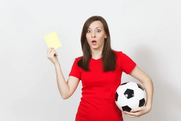 European serious severe young woman, football referee or player in red uniform showing yellow card, holding soccer ball isolated on white background. Sport play football, healthy lifestyle concept.