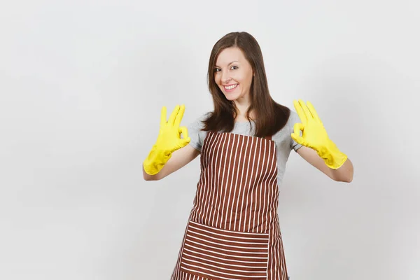 Junge attraktive lächelnde brünette Hausfrau in gestreifter Schürze, gelbe Handschuhe isoliert auf weißem Hintergrund. schöne Haushälterin zeigt ok Geste aussehende Kamera. Kopierfläche für Werbung. — Stockfoto
