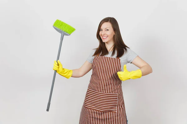 Young smiling housewife in striped apron, yellow gloves isolated on white background. Woman showing thumb up, cleaning maid holding sweeping with broom. Copy space for advertisement. Advertising area. — Stock Photo, Image