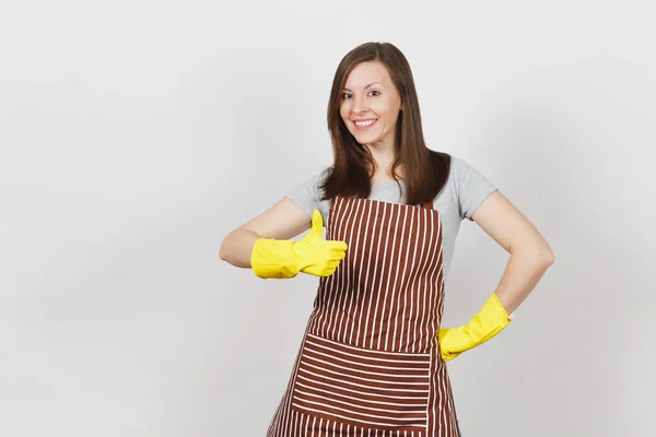 Joven atractiva y sonriente ama de casa caucásica en delantal rayado, guantes amarillos aislados sobre fondo blanco. Hermosa ama de llaves mostrando el pulgar hacia arriba mirando la cámara. Copiar espacio para publicidad . —  Fotos de Stock