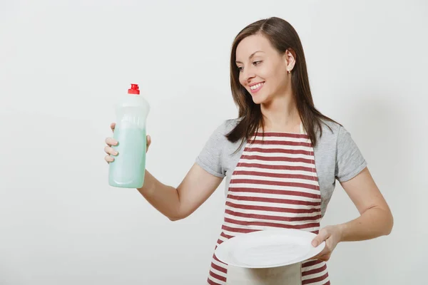 Jonge huisvrouw in gestreepte schort geïsoleerd op een witte achtergrond. Huishoudster vrouw houdt fles met schonere vloeistof voor wassen gerechten, witte lege ronde plaat. Fles met kopie ruimte voor reclame. — Stockfoto