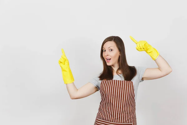 Young attractive smiling housewife in striped apron, yellow gloves isolated on white background. Beautiful housekeeper woman pointing index fingers aside looking camera. Copy space for advertisement. — Stock Photo, Image