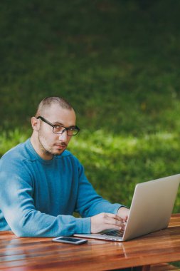 Genç başarılı adam işadamı veya öğrenci rahat mavi gömlekli, cep telefonu, dizüstü bilgisayar kullanarak şehir parkı ile masada oturan gözlük akıllı açık havada çalışan, kamera arıyorum. Mobil ofis kavramı.