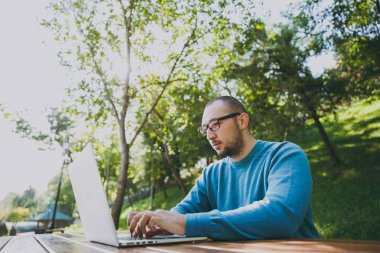 Genç başarılı akıllı adam işadamı veya öğrenci şehir Park açık havada yeşil doğa üzerinde çalışan dizüstü kullanarak cep telefonu ile masada oturan rahat mavi gömlekli gözlüklü. Mobil ofis kavramı.