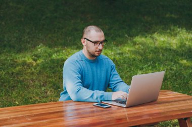 Genç başarılı akıllı adam işadamı veya öğrenci şehir Park açık havada yeşil arka plan üzerinde çalışan dizüstü kullanarak cep telefonu ile masada oturan rahat mavi gömlekli gözlüklü. Mobil ofis kavramı.