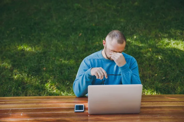 Young tired sad man businessman or student in casual blue shirt, glasses sitting at table with mobile phone in city park using laptop, working outdoors, worries about problems. Mobile Office concept.