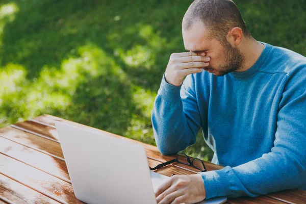 Giovane stanco uomo triste uomo d'affari o studente in camicia blu casual, occhiali seduti a tavola con il telefono cellulare nel parco della città utilizzando il computer portatile, lavorando all'aperto, preoccupazioni per i problemi. Concetto Mobile Office . — Foto Stock