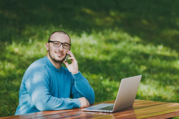 若い笑顔スマート男ビジネスマンやカジュアルな青いシャツを着て学生のテーブルに座っている眼鏡成功した屋外での作業、ラップトップを使用して都市公園で携帯電話で話しています。モバイル オフィス コンセプト. — ストック写真