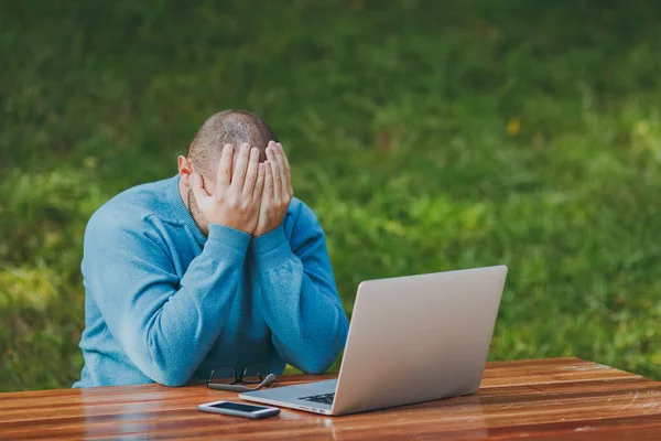 Üzgün kızgın şok adam işadamı veya öğrenci rahat mavi gömlekli, gözlük kapak yüz eller, sorunlar hakkında endişe ile şehir parkı kullanım dizüstü iş açık havada, masada otur. Mobil ofis kavramı.