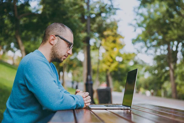 Giovane uomo intelligente di successo uomo d'affari o studente in camicia blu casual, occhiali seduti a tavola con il telefono cellulare nel parco della città utilizzando il computer portatile, lavorando all'aperto. Mobile Office concept. Vista laterale . — Foto Stock