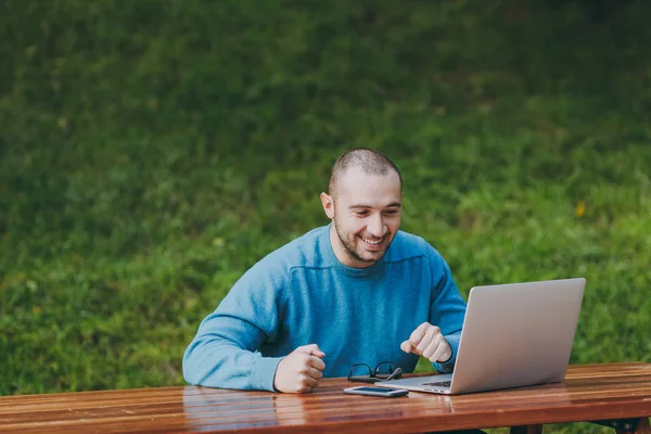 Genç başarılı mutlu akıllı adam işadamı ya da doğa dışarıda üzerinde çalışma öğrenci gözlük cep telefonu, dizüstü bilgisayar kullanarak şehir parkı ile masada oturan rahat mavi gömlekli. Mobil ofis kavramı.