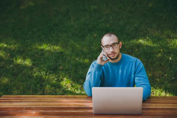 Young successful smiling smart man businessman or student in casual blue shirt, glasses sitting at table, talking on mobile phone in city park using laptop, working outdoors. Mobile Office concept.