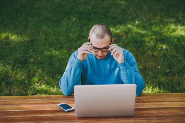 Jovem homem inteligente bem sucedido homem de negócios ou estudante em óculos casuais camisa azul sentado à mesa com telefone celular no parque da cidade usando laptop trabalhando ao ar livre na natureza verde. Conceito de escritório móvel . — Fotografia de Stock