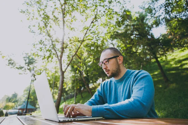 Genç başarılı akıllı adam işadamı veya öğrenci şehir Park açık havada yeşil doğa üzerinde çalışan dizüstü kullanarak cep telefonu ile masada oturan rahat mavi gömlekli gözlüklü. Mobil ofis kavramı.
