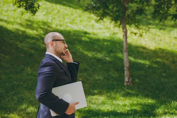 Успешный умный бизнесмен в белой рубашке, классическом костюме, очках. Мужчина стоит с ноутбуком компьютер, говорить по мобильному телефону в городском парке на открытом воздухе на фоне природы. Вид сбоку, бизнес-концепция . — стоковое фото