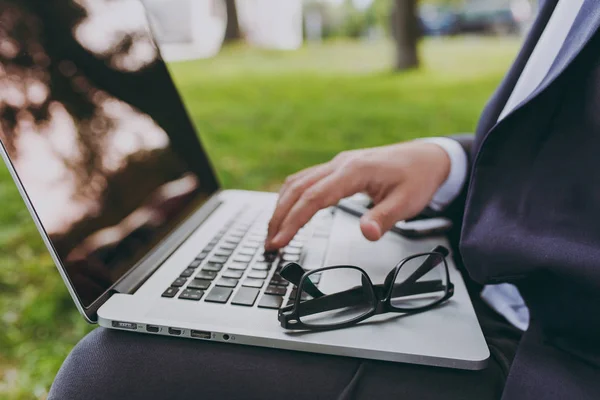 Close up cropped of hands on the keyboard. Businessman in classic suit, glasses. Man sit on soft pouf, work on laptop pc computer in city park on green lawn outdoors. Mobile Office concept. Side view.