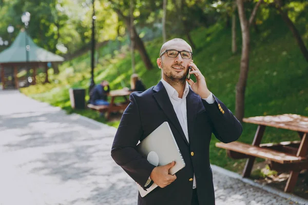 Успешный умный бизнесмен в белой рубашке, классическом костюме, очках. Мужчина стоит с ноутбуком компьютер, говорить по мобильному телефону в городском парке на открытом воздухе на фоне природы. Мобильный офис, концепция бизнеса . — стоковое фото