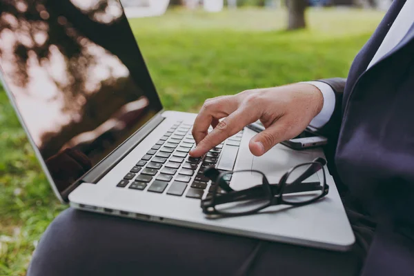 Close up cropped of hands on the keyboard. Businessman in classic suit, glasses. Man sit on soft pouf, work on laptop pc computer in city park on green lawn outdoors. Mobile Office concept. Side view.