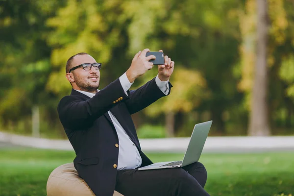 Młody biznesmen w klasyczny garnitur, biała koszula, okulary. Człowiek siedzieć na miękką pufę z komputerem pc laptop, robić selfie na telefon komórkowy w parku miejskim na zielony trawnik na zewnątrz. Koncepcja biznesowa Mobile Office. — Zdjęcie stockowe