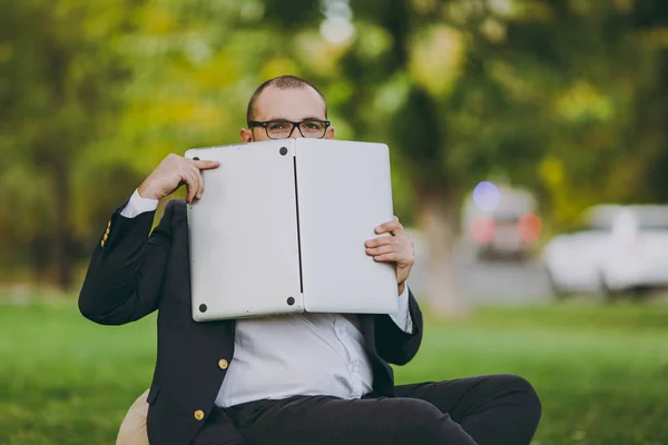 Jovem empresário de sucesso em camisa branca, terno clássico, óculos. Homem sentar-se em pufe macio, esconder-se atrás do computador portátil pc no parque da cidade no gramado verde ao ar livre na natureza. Escritório móvel, conceito de negócio . — Fotografia de Stock