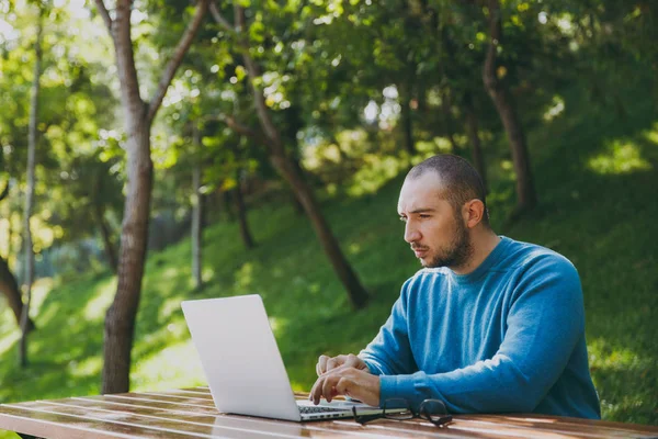 Genç başarılı akıllı adam işadamı veya öğrenci şehir Park açık havada yeşil doğa üzerinde çalışan dizüstü kullanarak cep telefonu ile masada oturan rahat mavi gömlekli gözlüklü. Mobil ofis kavramı.