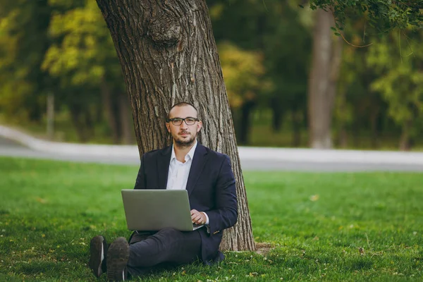 Giovane uomo d'affari di successo in camicia bianca, abito classico, occhiali. L'uomo siede su terreno erboso, lavora su computer di computer di computer portatile in parco di città su prato verde all'aperto su natura. Mobile Office, concetto di business . — Foto Stock