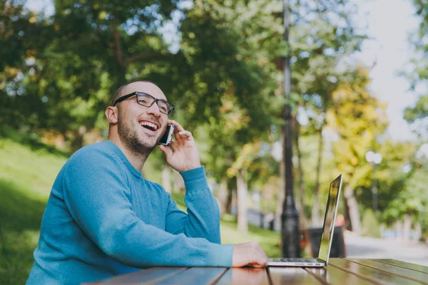 Молодой успешный улыбающийся умный бизнесмен или студент в повседневной синей рубашке, в очках, сидящих за столом, разговаривающих по мобильному телефону в городском парке с помощью ноутбука, работающих на открытом воздухе. Концепция мобильного офиса . — стоковое фото