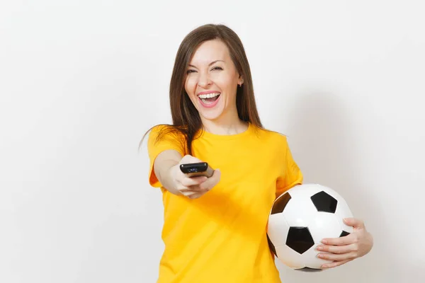 Beautiful European young cheerful woman, football fan or player in yellow uniform hold TV remote, soccer ball isolated on white background. Sport, play football, health, healthy lifestyle concept.