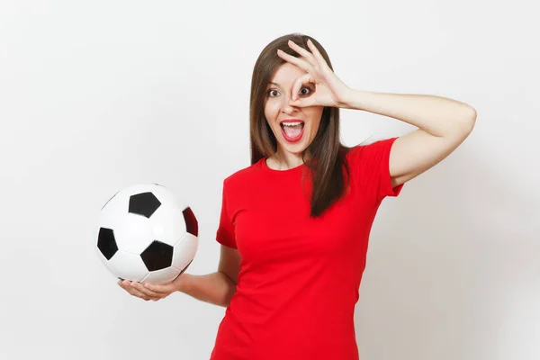 Pretty European young woman, football fan or player in red uniform holding classic soccer ball, fingers near eye isolated on white background. Sport, play football, health, healthy lifestyle concept.