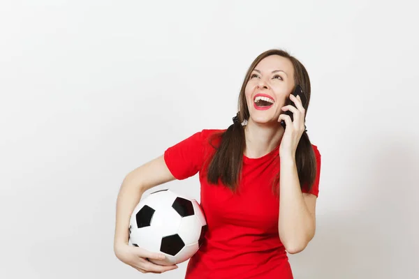 European young woman, two fun pony tails, football fan or player in red uniform hold soccer ball, talk on mobile phone isolated on white background. Sport play football, healthy lifestyle concept.